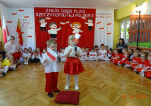 Na tle biało-czerwonej dekoracji stoi chłopiec z dziewczynką. Dzieci ubrane są na biało-czarwono. W rękach trzymają biało-czerwone chorągiewki i recytują wiersz. i